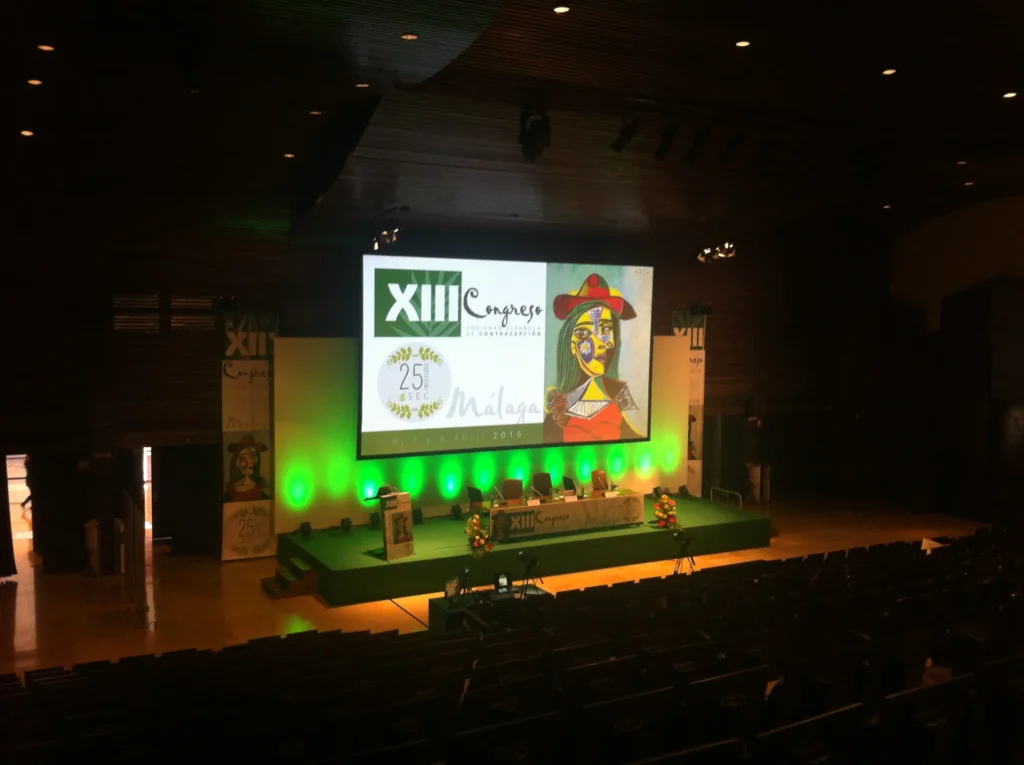 Audiovisual rental setup for a congress in Malaga, including a big screen, projector, LED lighting, podium, and sound system.