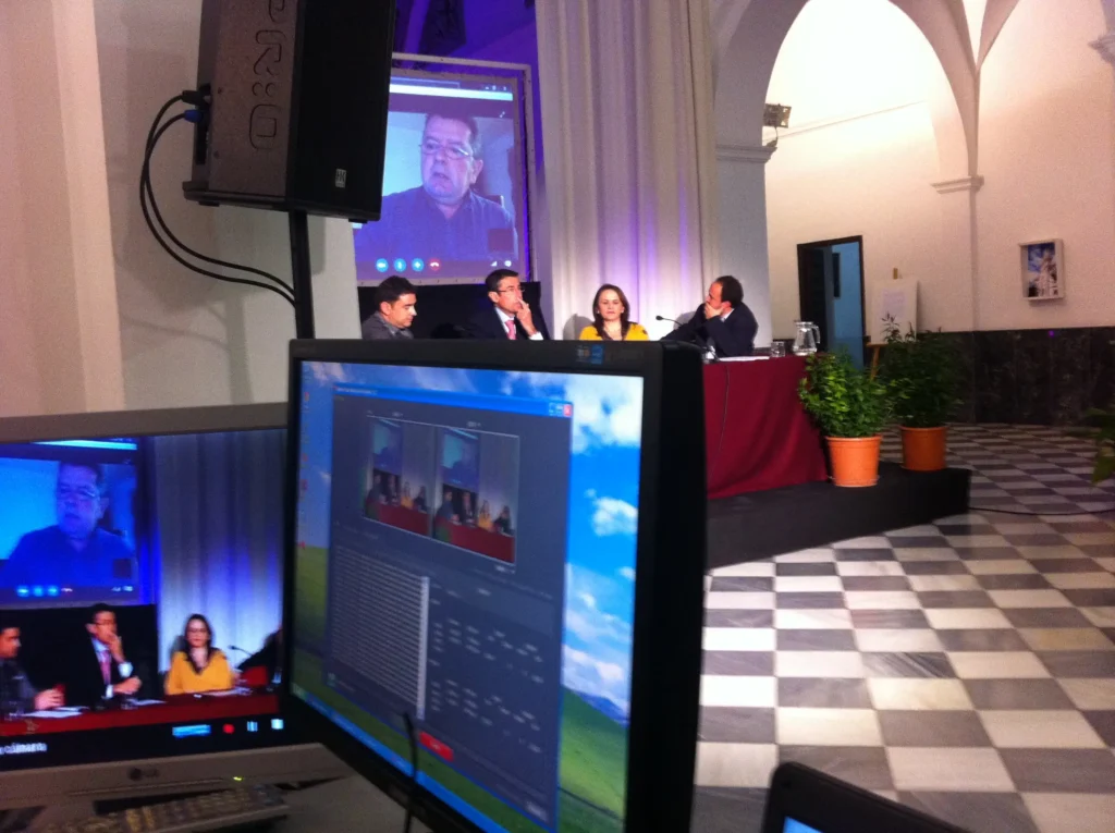 Technician operating the video-conferencing and camera control station for a hybrid panel event in Malaga, with a remote speaker shown live on the main screen.