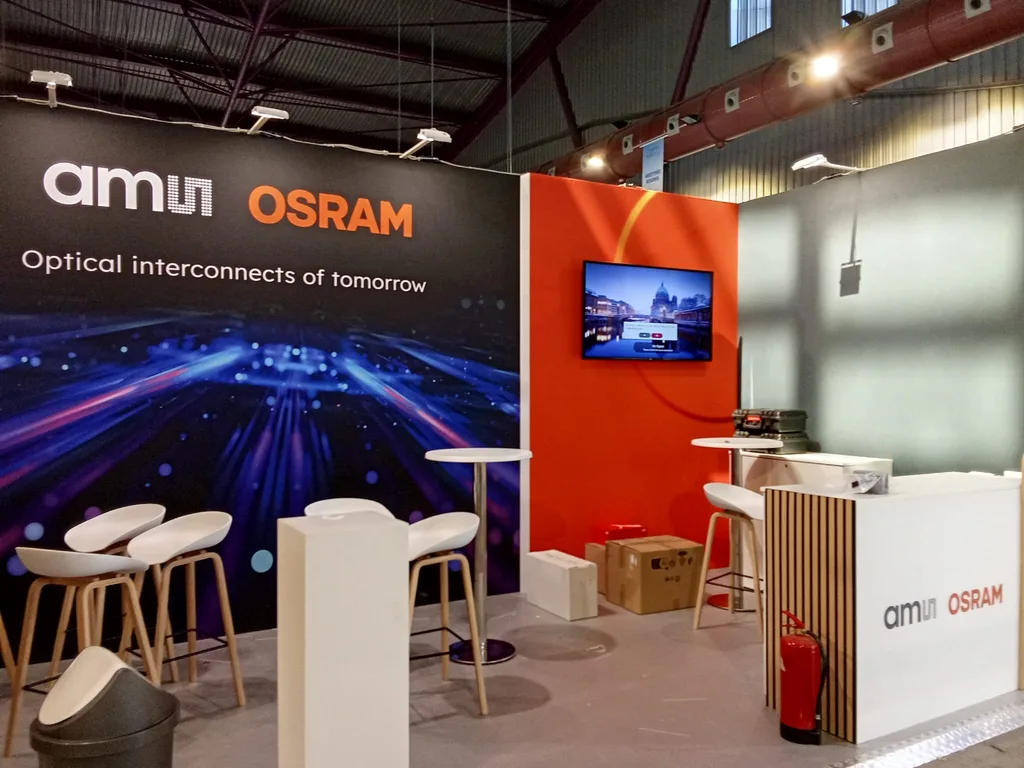 A single screen mounted on an orange stand wall at the ECOC 2026 exhibition, with bar stools and a reception counter already in place.
