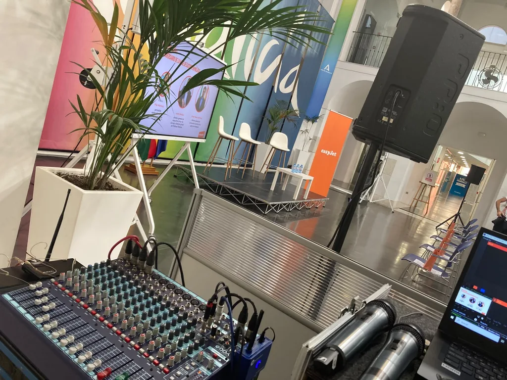 The sound position at the Tinkle event for easyJet, with a view of the stage. The sound desk with a mixing console, two handheld microphones and a laptop in the foreground, and a PA speaker on a stand and a low stage with a slide screen in the distance, at the Tinkle event for easyJet in Malaga.