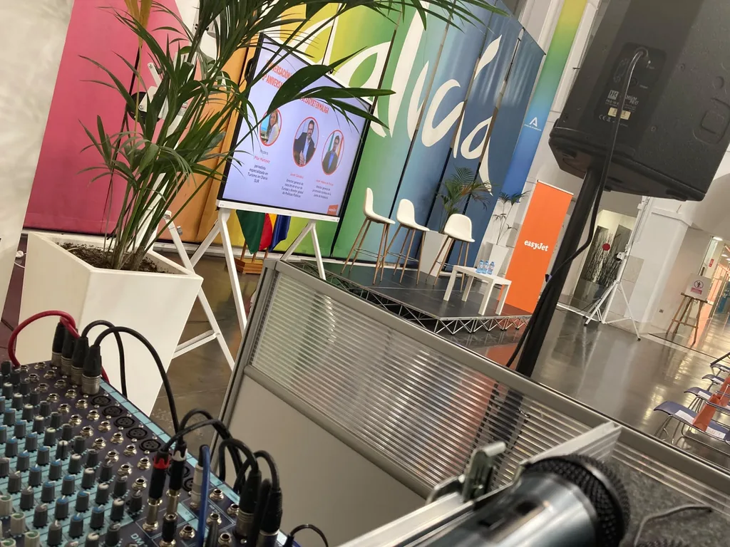 A handheld microphone ready beside the mixing console at the Tinkle event for easyJet in Malaga. A close view of a mixing console with cables plugged in and a handheld microphone beside it, with a PA speaker on a stand and a slide screen out of focus behind, at the Tinkle event for easyJet in Malaga.