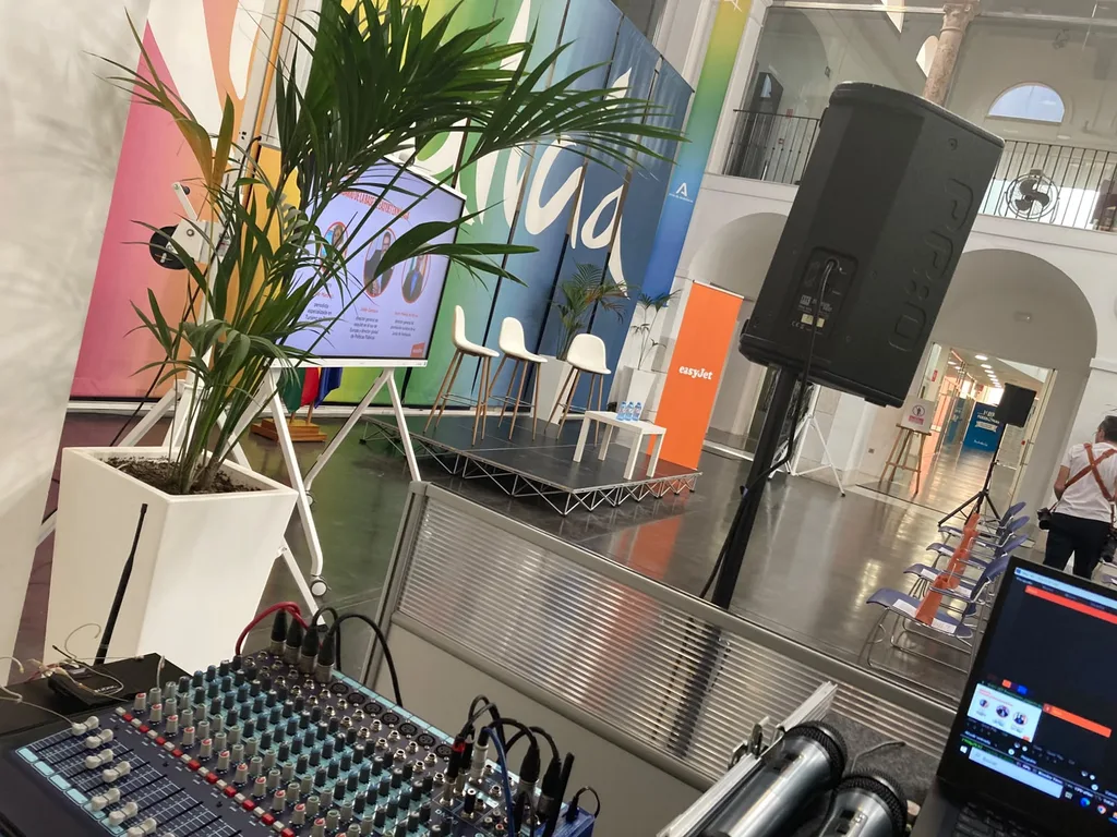 The sound desk and the sound system we supplied for the Tinkle event for easyJet, with the stage and the screen behind, which are not ours. A mixing console and two handheld microphones next to a laptop, with a PA speaker on a stand and a low stage with a slide screen behind, at the Tinkle event for easyJet in Malaga.