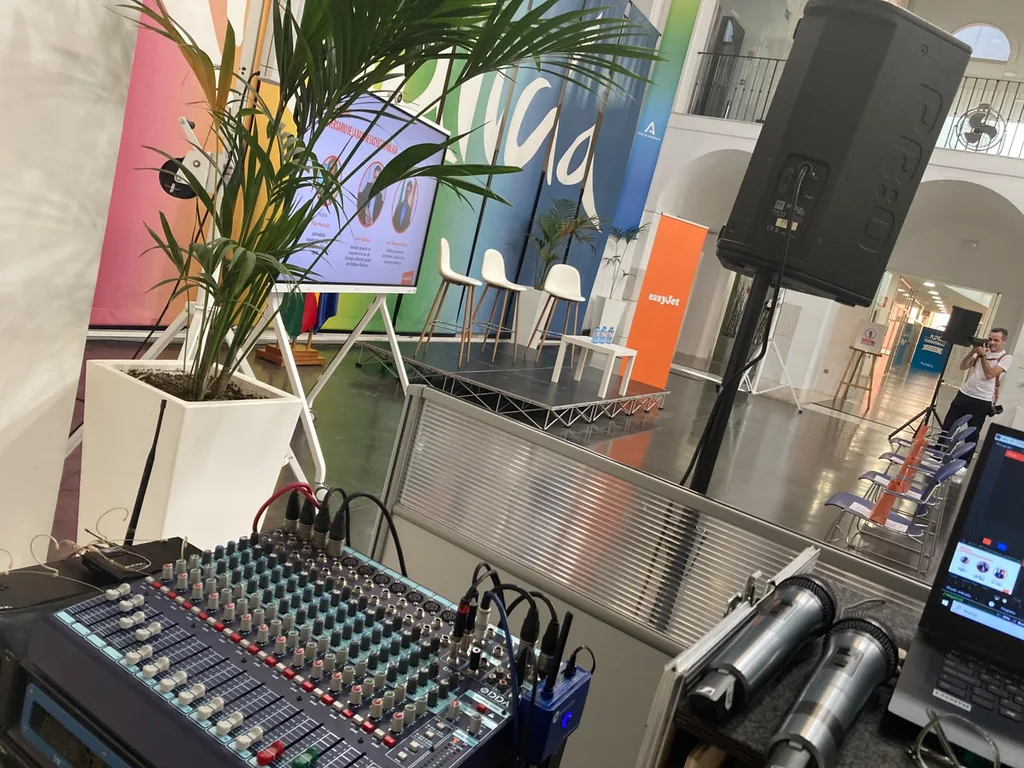 Malaga Event Gear supplied the sound system and a technician for this Tinkle event for easyJet in the centre of Malaga on 22 October 2026. A mixing console with two handheld microphones in the foreground and a PA speaker on a stand, facing a low stage with a screen and seats, at an event for Tinkle and easyJet in the centre of Malaga.